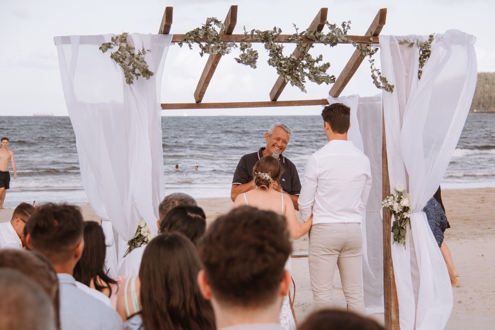 Couple exchanging vows under a draped arch on a beach as guests watch, the kind of port-of-call ceremony a destination wedding cruise can include