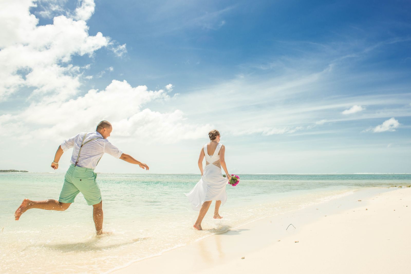 Bride and groom running through shallow turquoise water on a sunny beach after their wedding