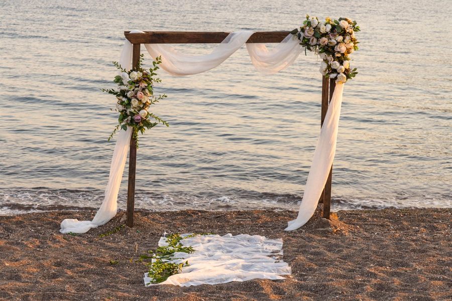 Wooden wedding arch draped with white fabric and flowers on a beach at sunset
