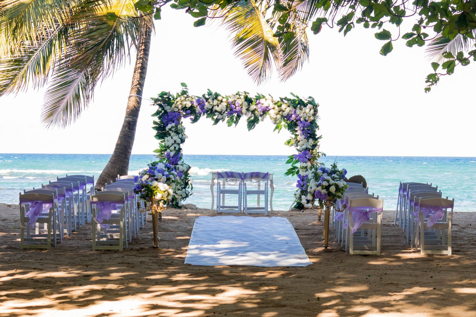 Floral wedding arch and white chairs on a palm-shaded beach, a setting some wedding cruise packages offer in port